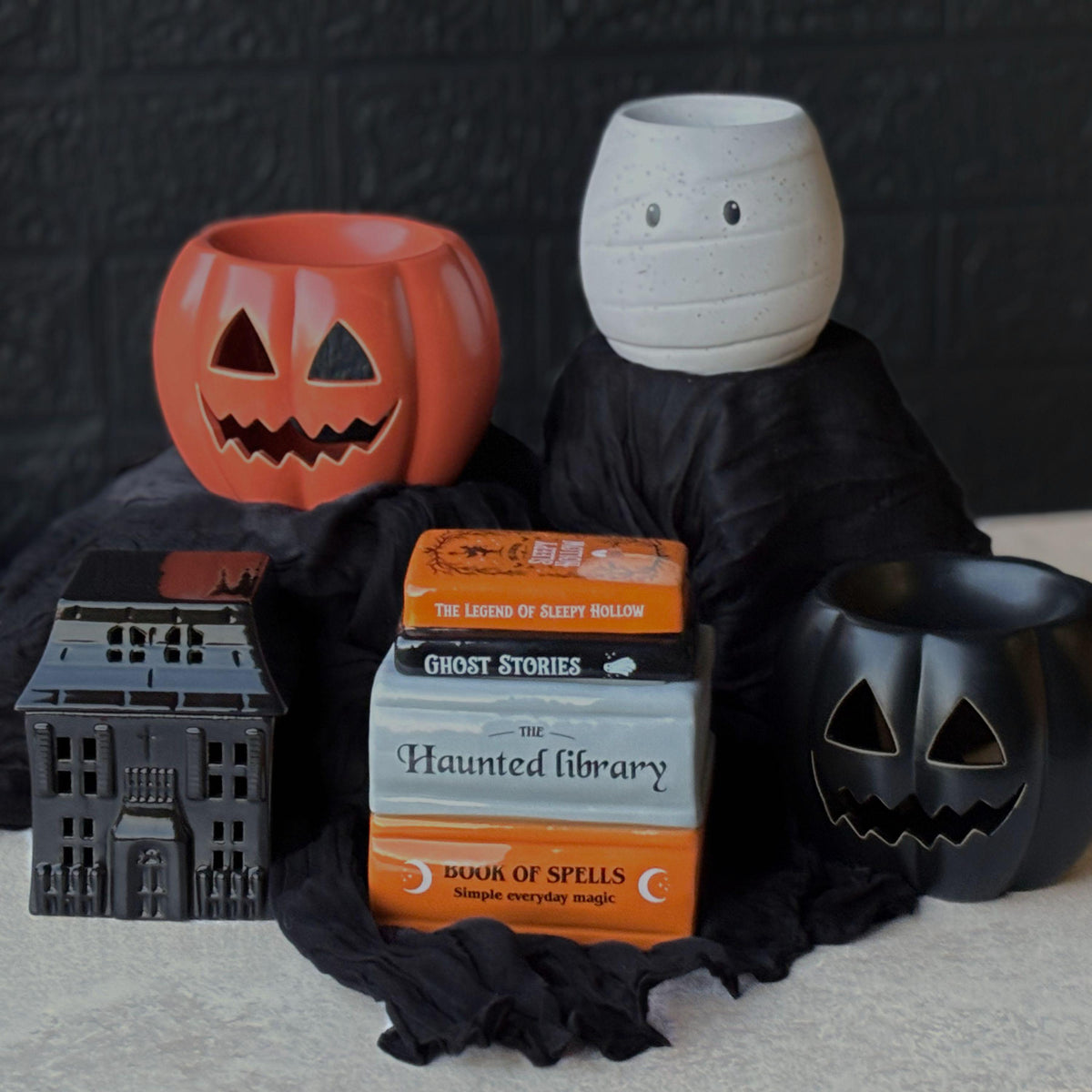 Halloween ceramic wax melt burners displayed together, including an orange pumpkin, black pumpkin, white mummy, haunted house and stack of Halloween books, styled with black fabric for a spooky autumn home décor scene.  Halloween Wax Melts
Hug In A Box Gifts
Boo Basket Gift Idea
Home Fragrance Gift
Halloween Wax Burner
Wax Melt Burners
Seasonal Oil Burners
Witchy Home Decor
Gothic Melt Burner
Ceramic Wax Burners
Pumpkin Wax Burner
Jack O Lantern
Spooky haunted House